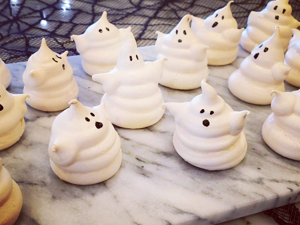 White ghost-shaped meringue cookies with candy eyes, fully dried and crisp, arranged on plate.