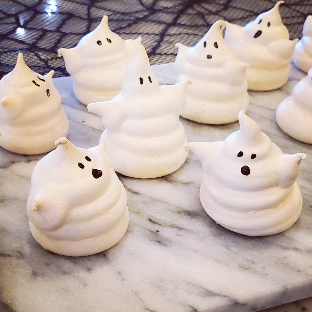 White ghost-shaped meringue cookies with candy eyes, fully dried and crisp, arranged on plate.