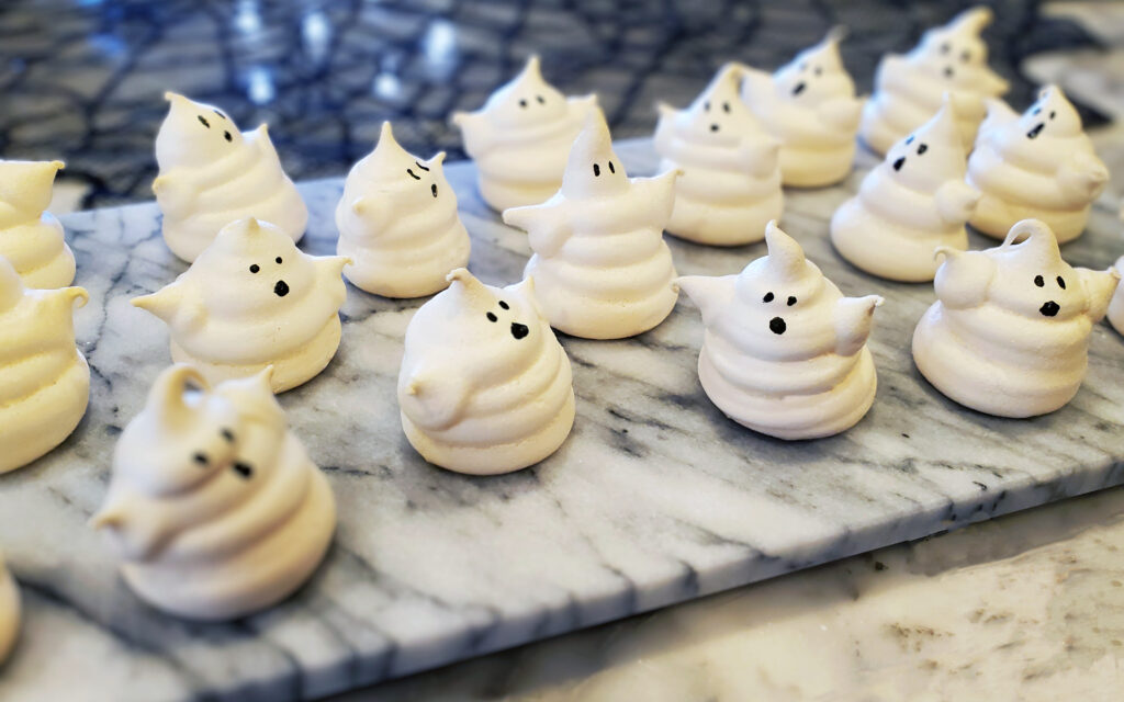 White ghost-shaped meringue cookies with candy eyes, fully dried and crisp, arranged on plate.