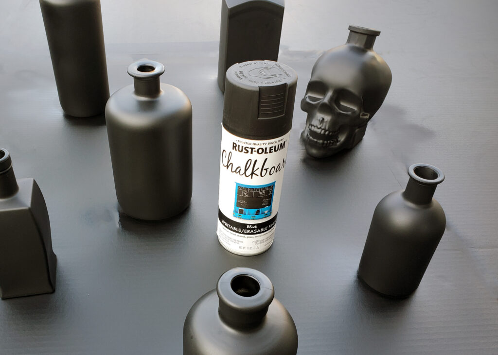 Glass bottles coated in matte black chalkboard spray paint, drying between coats for DIY potion bottle décor.