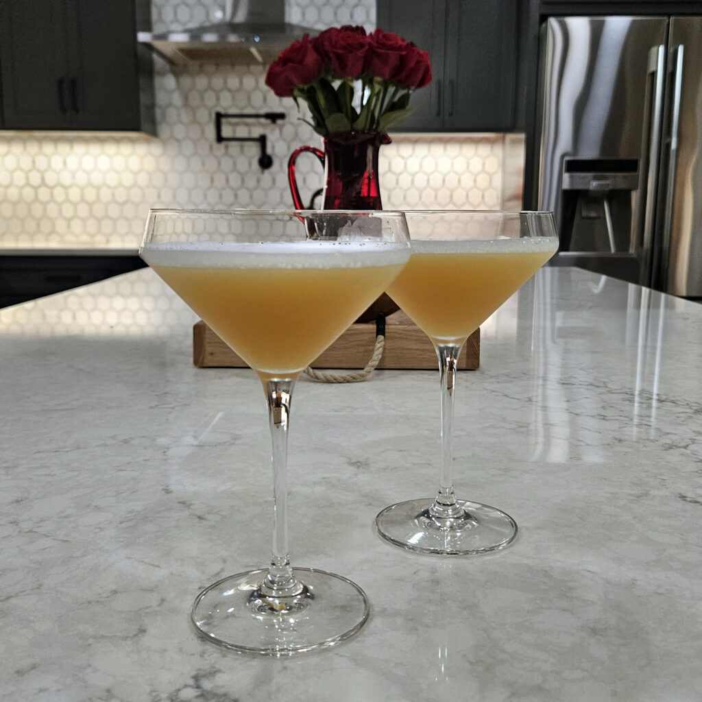 Two pale-gold Lola Sour cocktails served in coupe glasses on a marble kitchen island, topped with a light frothy foam. A vase of red roses and a modern kitchen backdrop add a romantic, polished feel to the cocktail presentation.