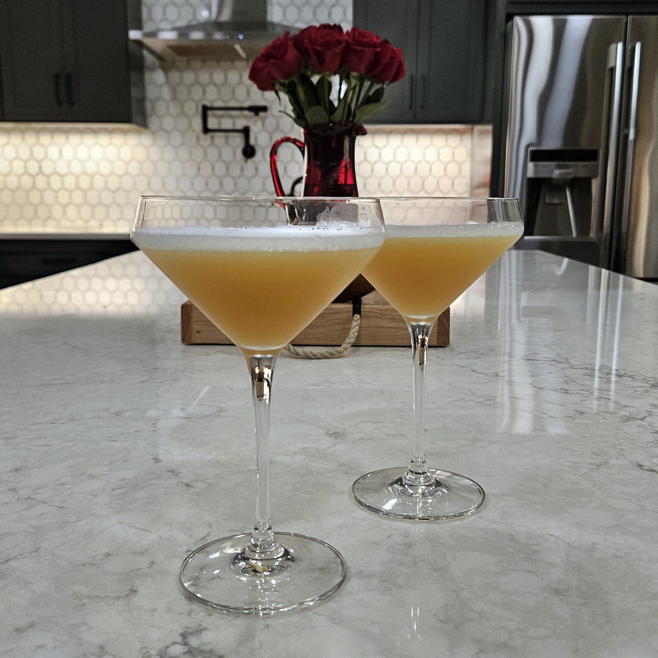 Two pale-gold Lola Sour cocktails served in coupe glasses on a marble kitchen island, topped with a light frothy foam. A vase of red roses and a modern kitchen backdrop add a romantic, polished feel to the cocktail presentation.