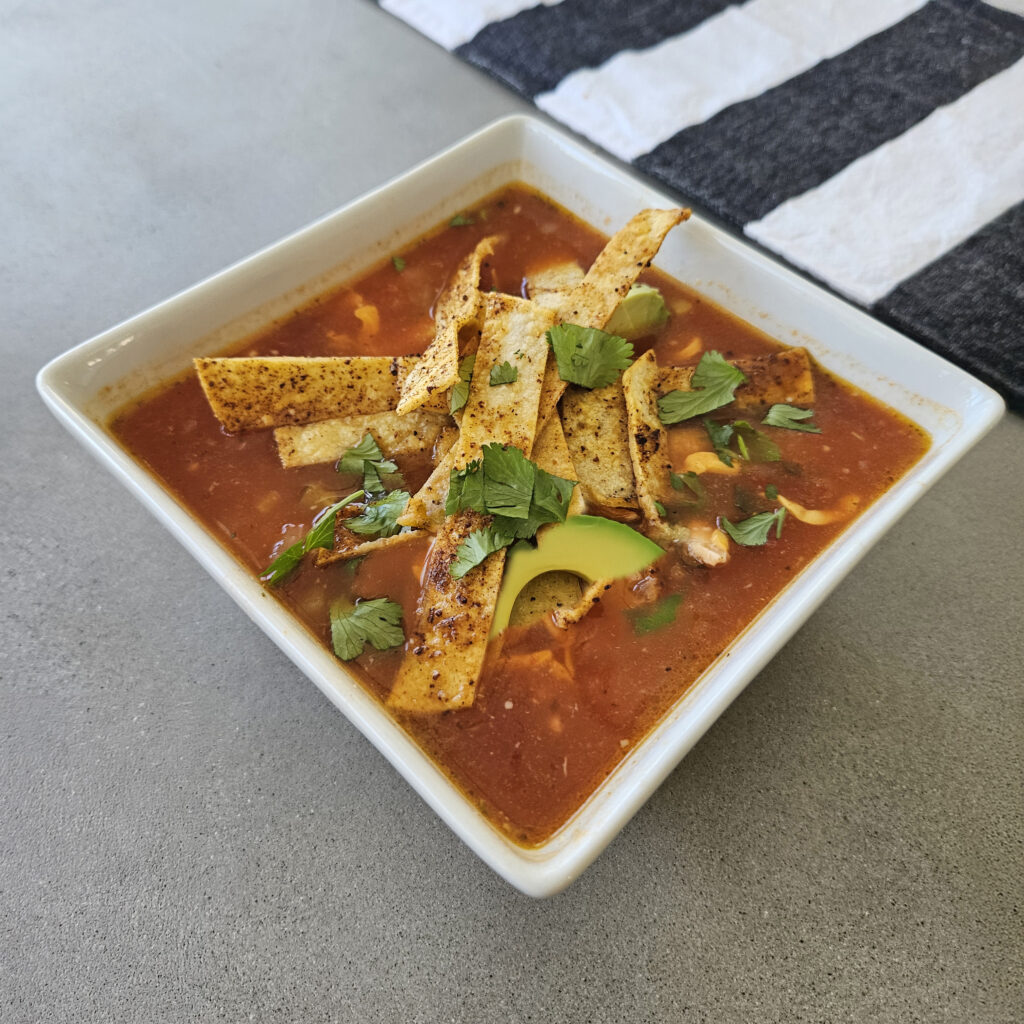 Finished soup topped with avocado, cheese, tortilla strips, cilantro, and lime.