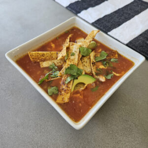 Finished soup topped with avocado, cheese, tortilla strips, cilantro, and lime.
