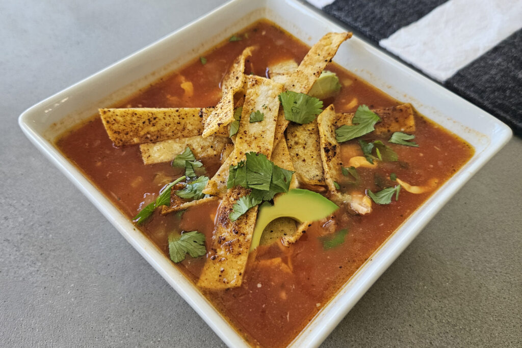 Finished soup topped with avocado, cheese, tortilla strips, cilantro, and lime.