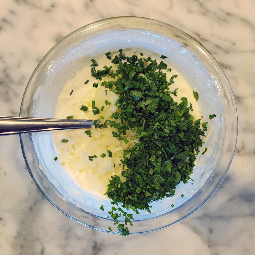 Fresh herbs, cream cheese, sour cream, and chopped onions prepared for making Green Goddess dip.