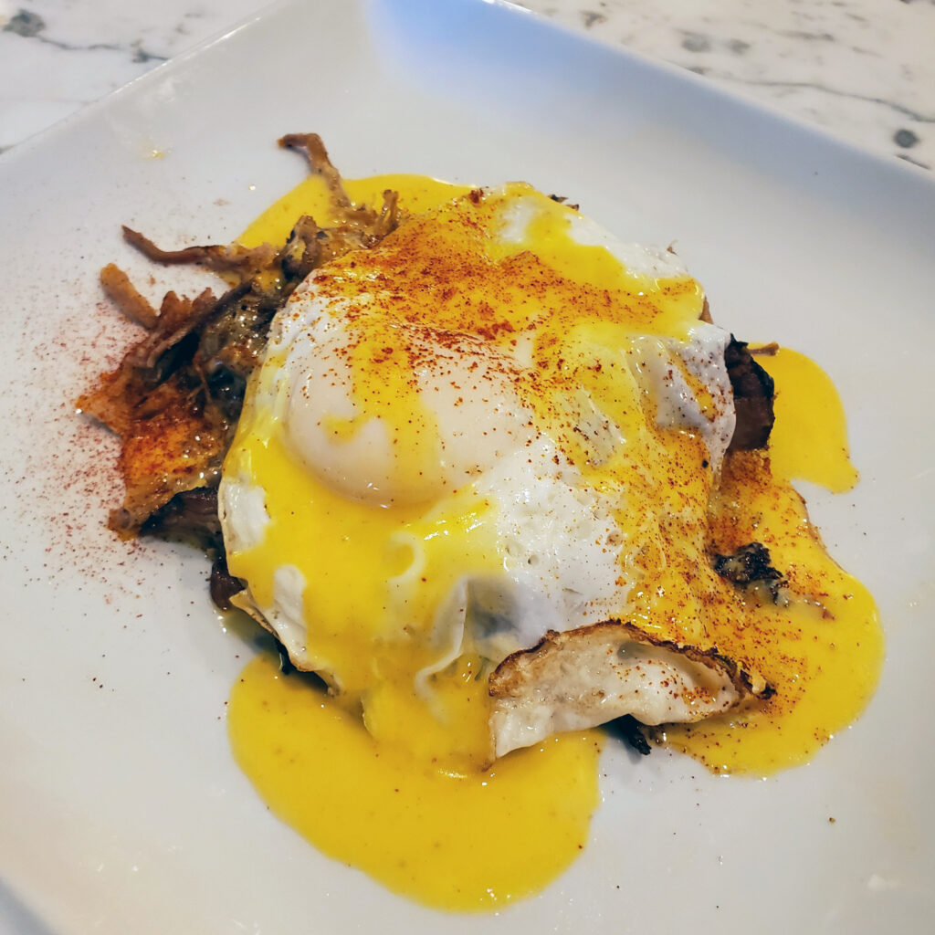 Crispy fingerling potato hash topped with smoked pulled pork, a sunny-side-up egg, and lemon hollandaise on a neutral plate.