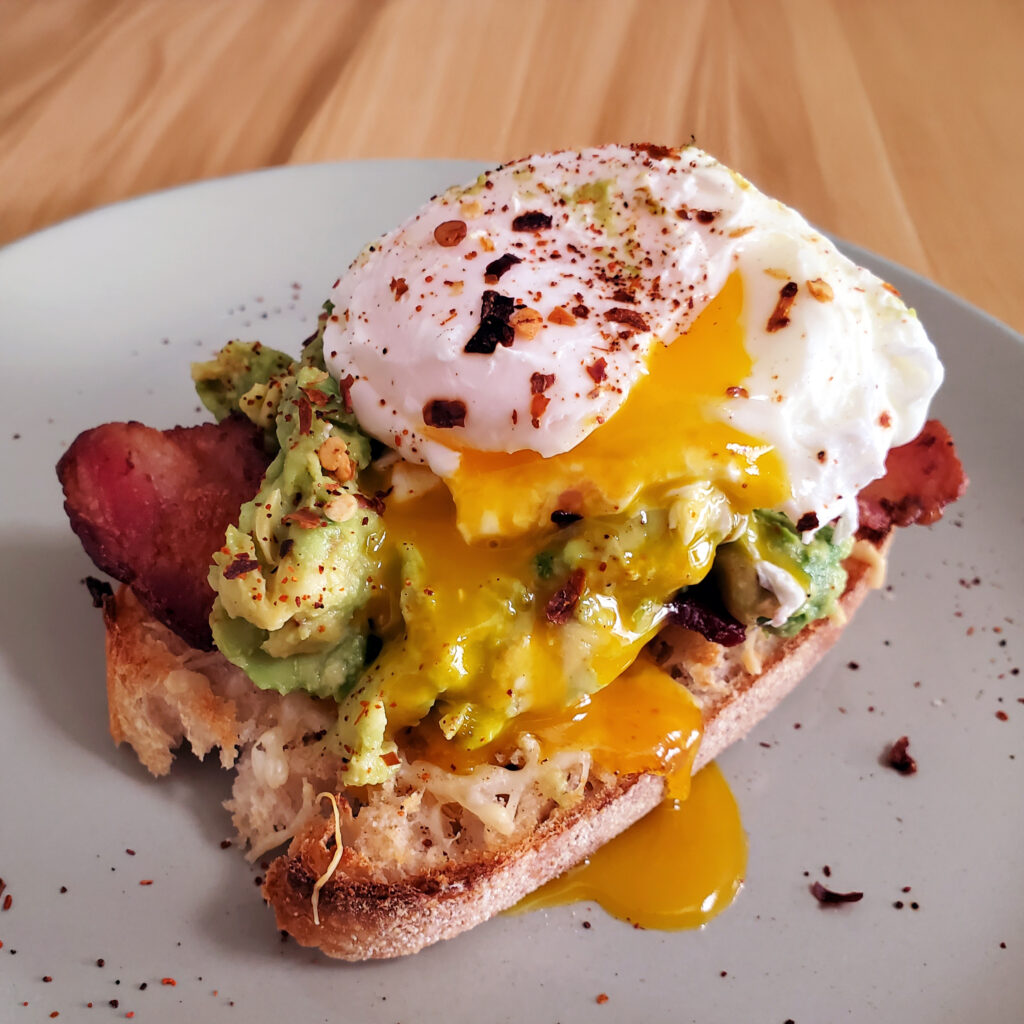 Avocado toast on crusty sourdough topped with crispy bacon, a poached egg, and red pepper flakes