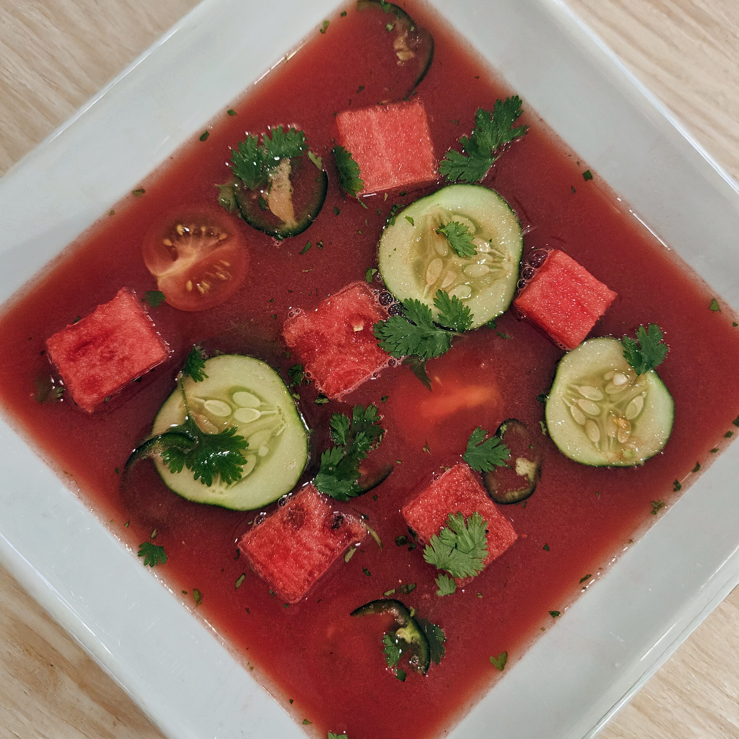 Chilled watermelon gazpacho served in a shallow white bowl, garnished with cucumber slices, watermelon cubes, cherry tomatoes, cilantro, and jalapeño slices.