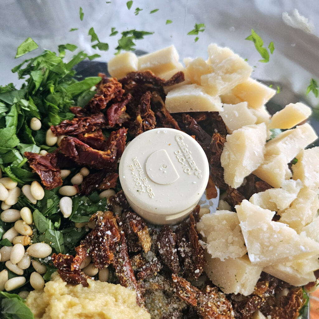 Food processor bowl filled with fresh basil leaves, sun-dried tomatoes, pine nuts, garlic, chunks of Parmigiano-Reggiano, and olive oil before blending into pesto.