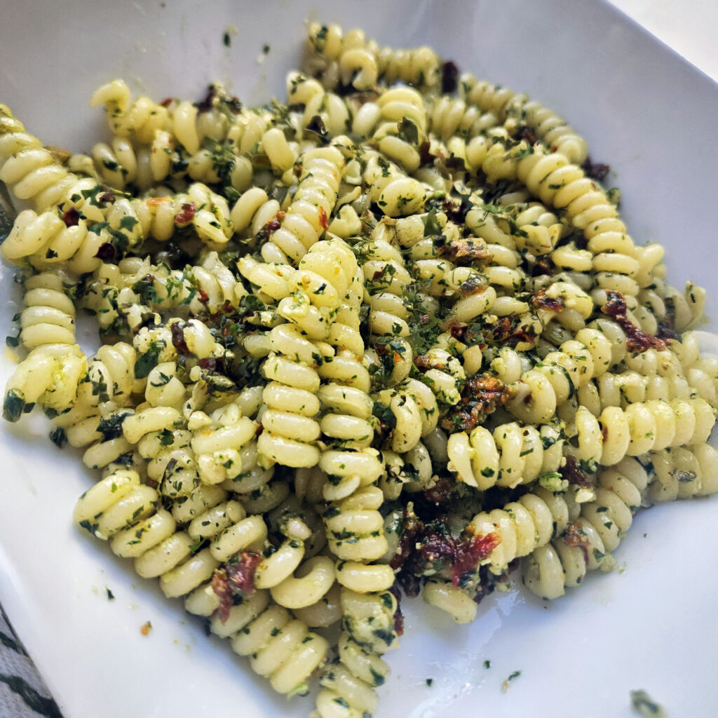 Sun-dried tomato pesto tossed with short, ridged pasta, showing flecks of basil, grated Parmigiano-Reggiano, and pieces of sun-dried tomato in a white serving bowl.